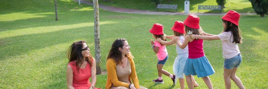 Happy children playing in a sunny park with two mothers sitting nearby, enjoying the outdoors.